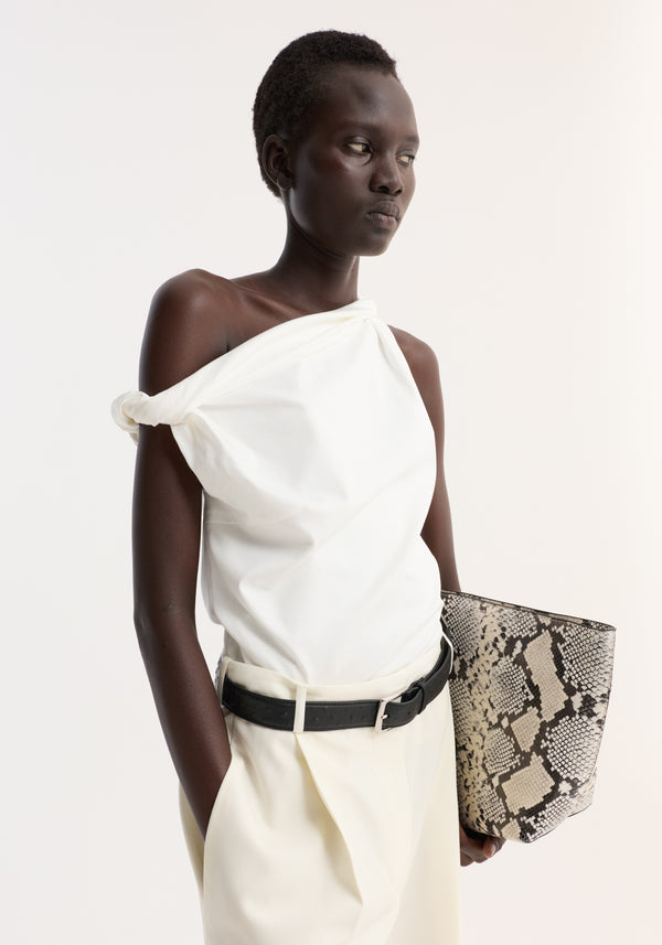 Dark-skinned model wearing the Sculptural cotton silk top, off white, with twisted strap detail, styled asymmetrically, paired with cream trousers and carrying a snakeskin tote.