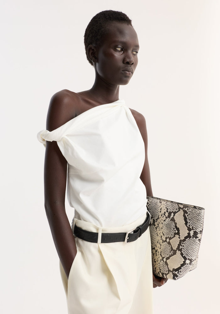 A woman models the Sculptural cotton silk top, featuring a sleeveless, twisted strap detail and asymmetrical drape, paired with high-waisted trousers and a snakeskin print bag.