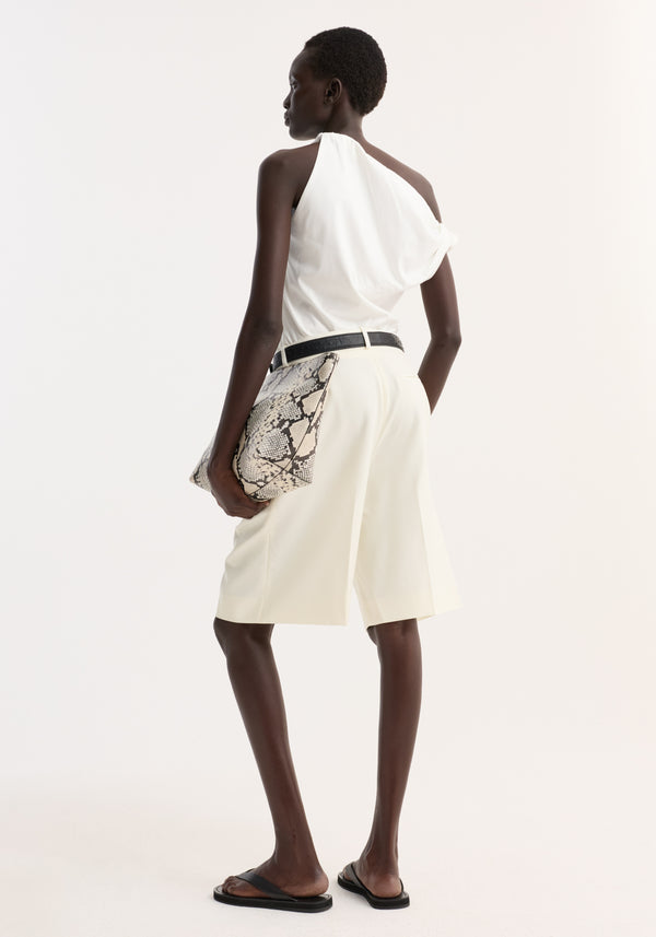 Model wearing Sculptural cotton silk top with twisted strap, asymmetrical drape, paired with high-waisted cream shorts, black belt, sandals, and holding a snakeskin-pattern clutch.