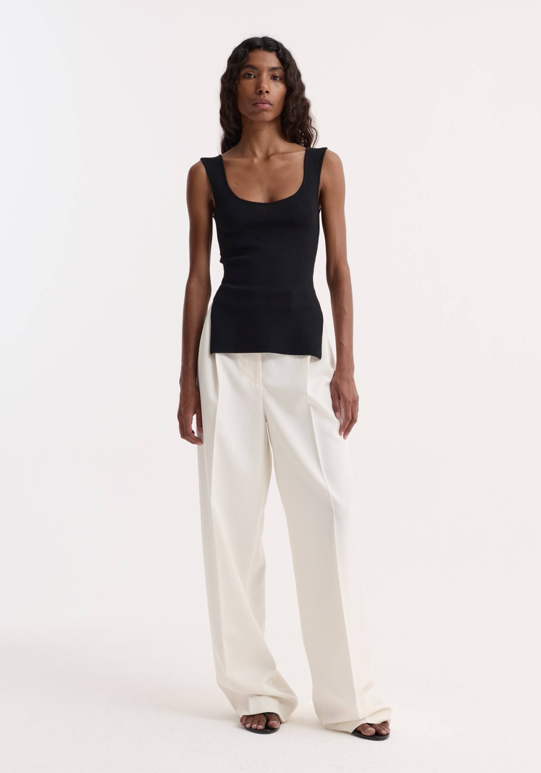 Model wearing Long knitted bustier top | black, with a scoop neckline and side slits, paired with high-waisted white trousers and black sandals.