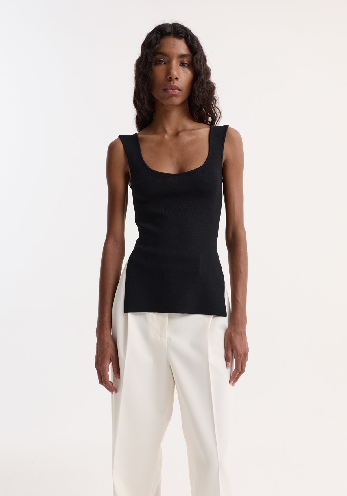 Woman wearing a Long knitted bustier top with scoop neckline, fitted design, and side slits, paired with high-waisted, loose-leg trousers, standing forward.