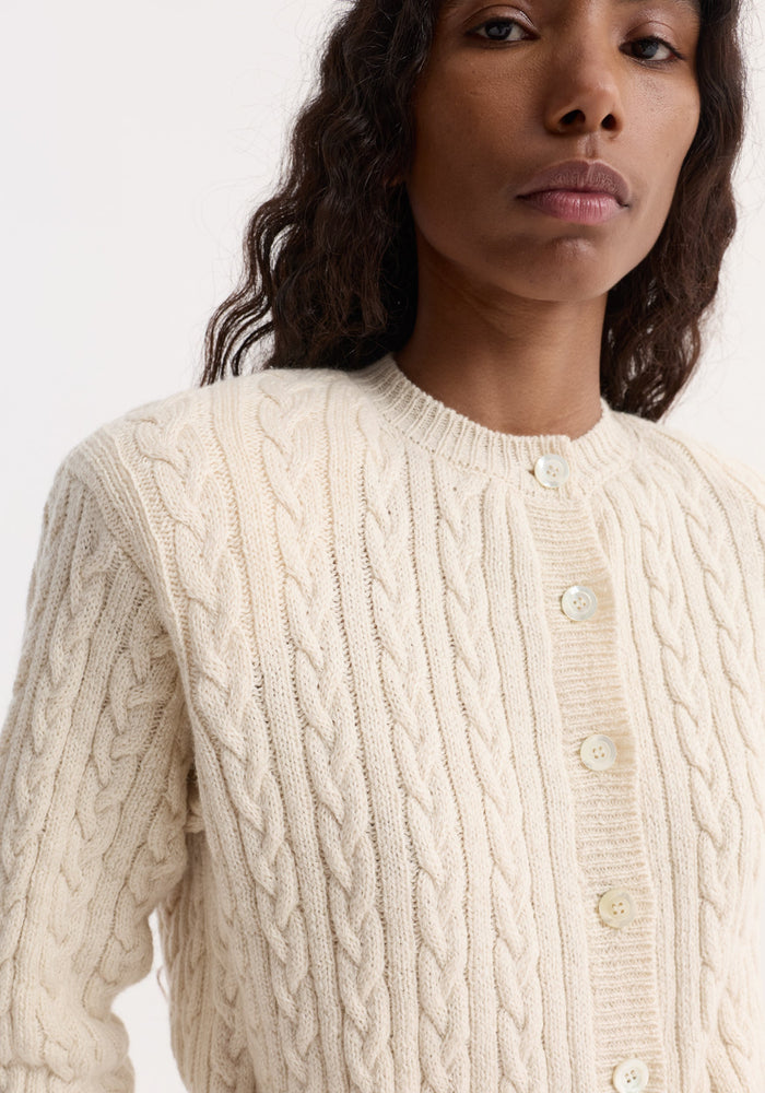 Fine merino cable cardigan in cream, featuring a classic braided pattern, subtle shoulder pads, and a slightly cropped fit, worn by a model.
