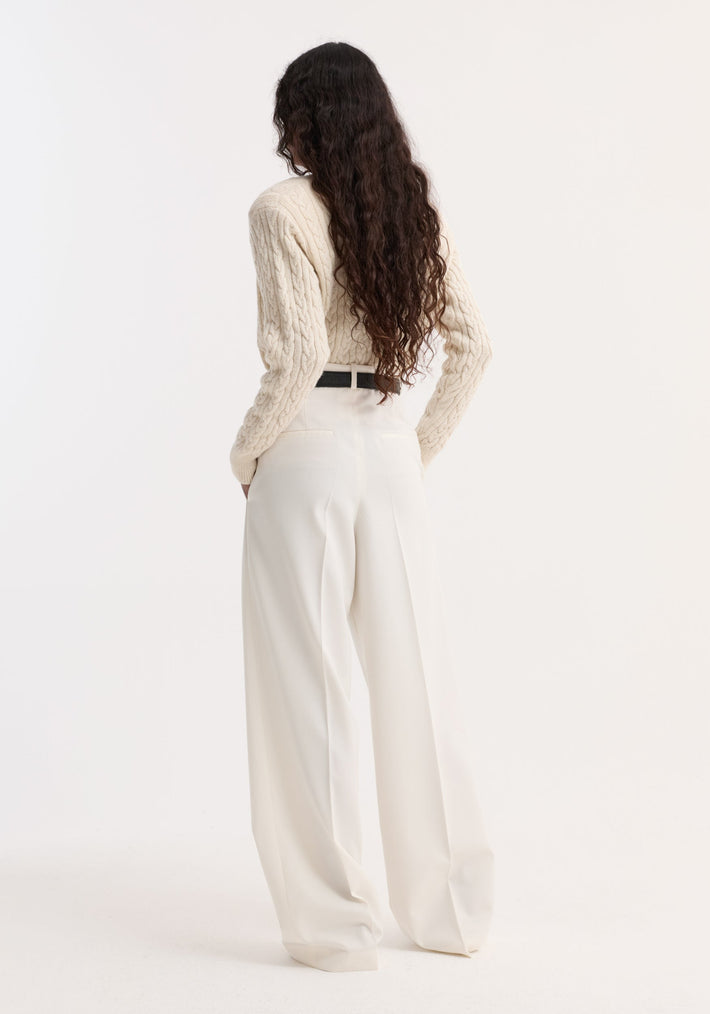 Rear view of a model wearing the Fine merino cable cardigan in cream, showcasing its slightly cropped fit and subtle shoulder pads, paired with high-waisted trousers.