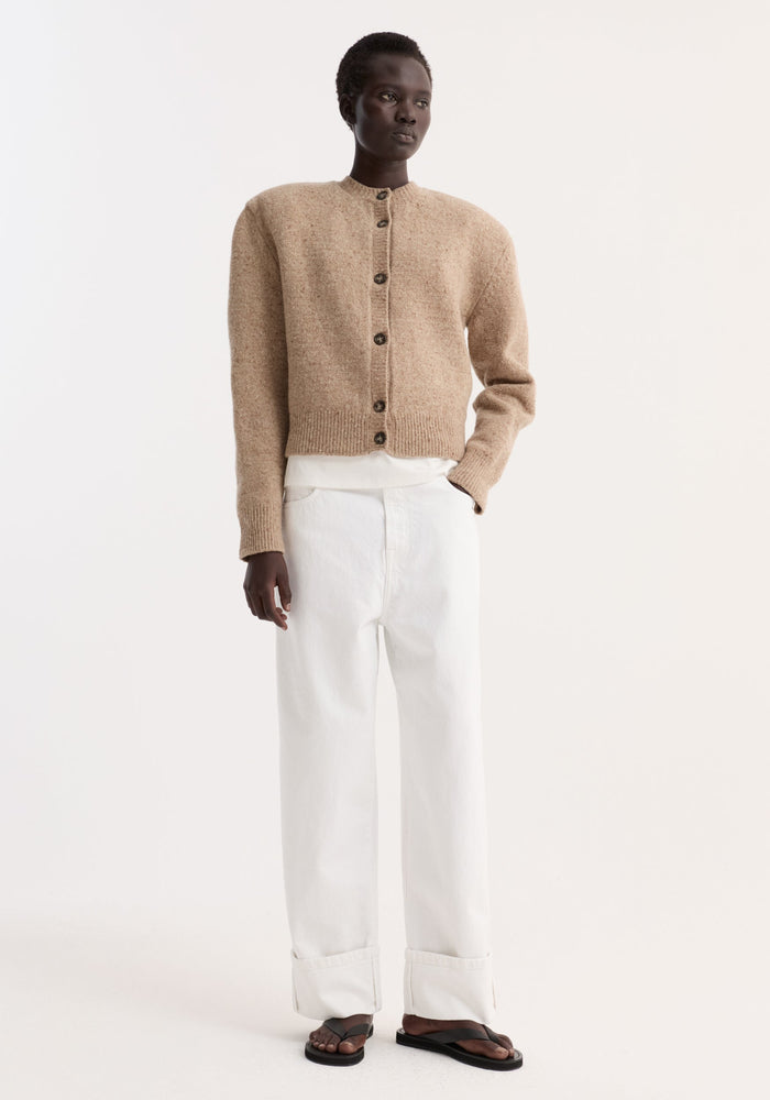 Woman in Fine merino tweed cardigan, oat melange, cropped with shoulder pads, over white top, paired with loose white trousers and black sandals.