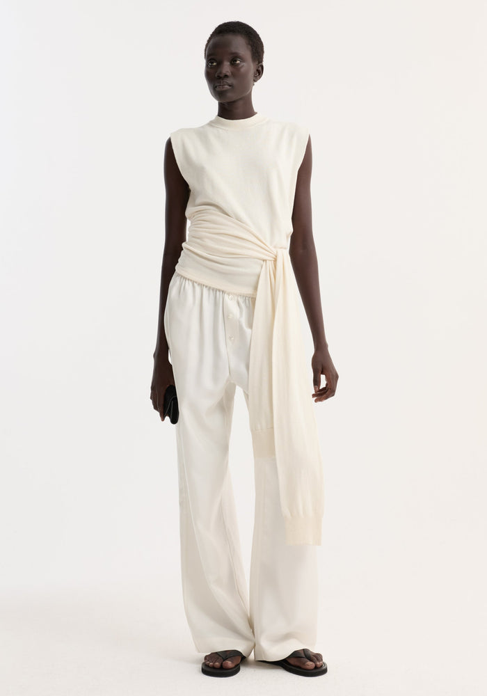 Model wearing an Open back knitted wrap top in off white, featuring a tied waist and open back, paired with wide-leg trousers and black sandals.