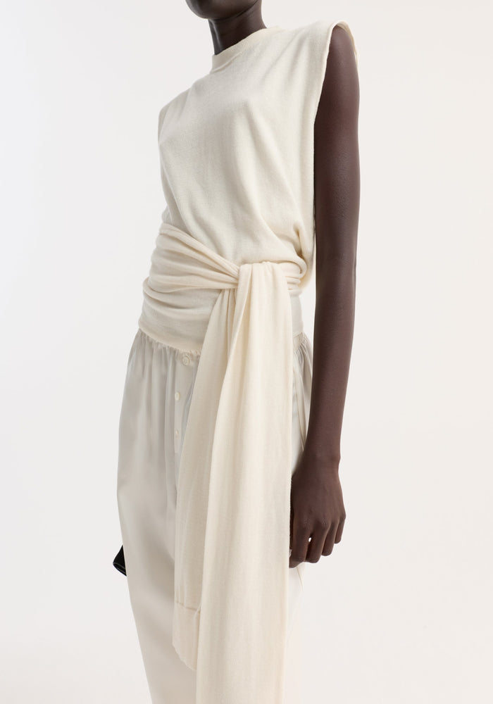 Model wearing Open back knitted wrap top in off white, featuring a crew neckline, wrap waist, and sash-style extensions, paired with button-detailed, loose-fitting trousers.