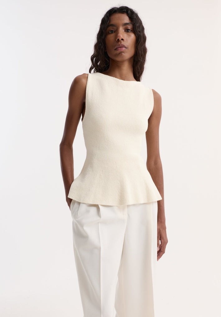 Woman wearing the Sculptural knitted top | cream, featuring a boat neckline, sleeveless design, and subtle peplum flare, paired with loose-fit trousers.