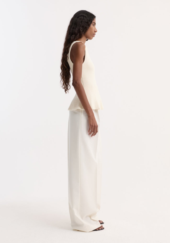 Model in profile wearing Sculptural knitted top | cream, featuring a sleeveless design with boat neckline and peplum flare, paired with high-waisted wide-leg trousers.