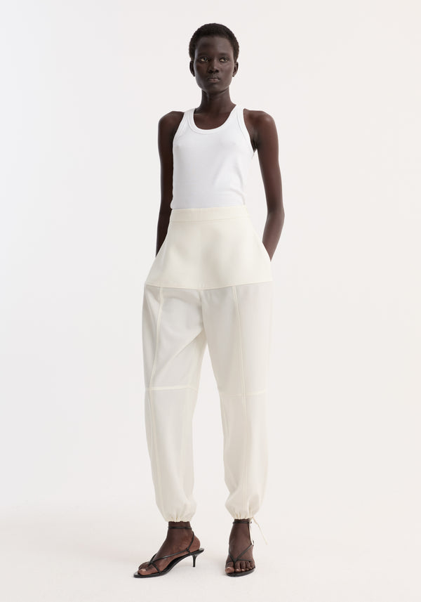 Dark-skinned model wearing Architectural trousers, featuring a fitted waist, seam detailing, elasticized cuffs, and adjustable hems, paired with a white ribbed tank top.