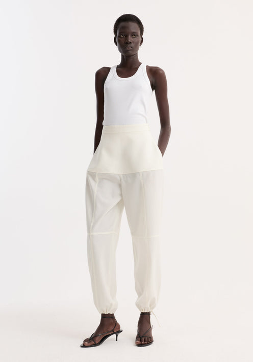Model wearing Sculptural trousers in off white, featuring a fitted waist, seam detailing, and adjustable hems, paired with a white tank top and black sandals.
