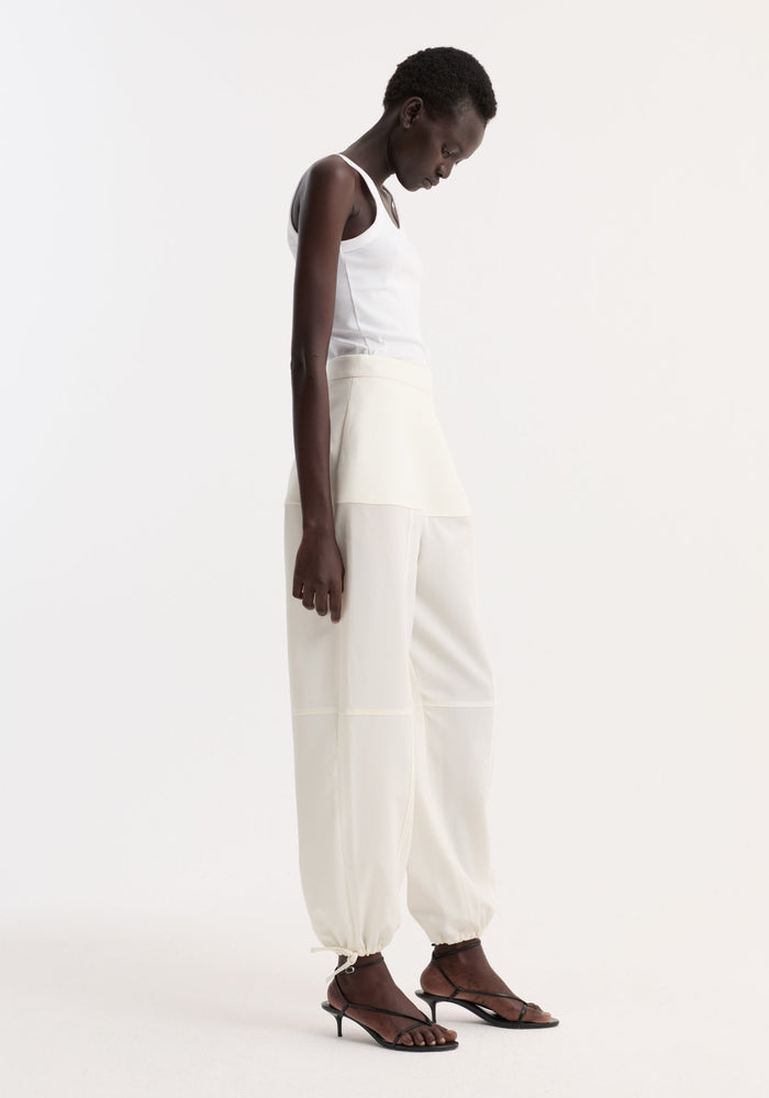 Model in profile wearing Sculptural trousers | off white, showcasing seam detailing and ankle adjusters, paired with a white ribbed tank top and black sandals.
