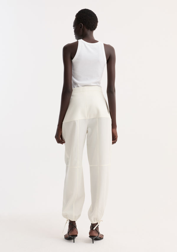 Back view of person wearing off-white Architectural trousers with seam detailing and ankle adjusters, paired with a white ribbed tank top and black sandals.