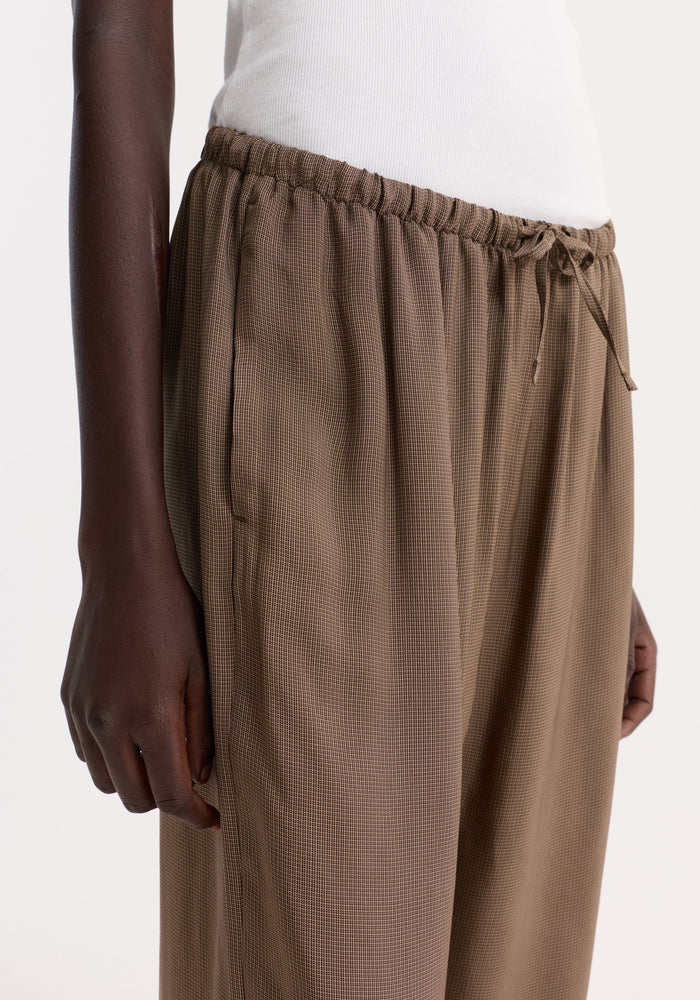 Person wearing relaxed fit trousers in brown mini check with a drawstring waistband and side pockets, paired with a white ribbed tank top.