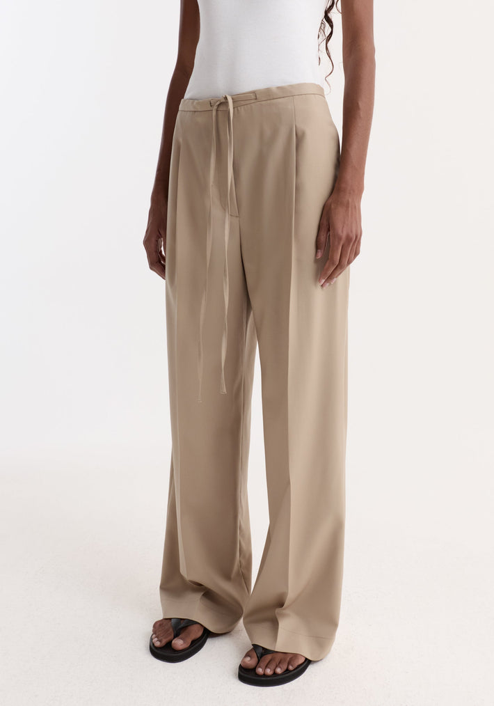Relaxed drawstring trousers in beige, featuring a wide-leg silhouette and elegant drawstring waistband, worn with black thong sandals and a white ribbed tank hem visible.