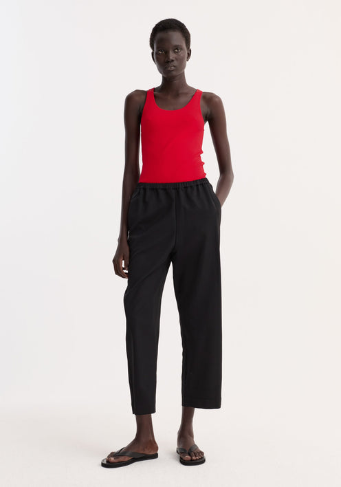 Model wearing cropped wide leg trousers in black, featuring an ankle-length fit, elasticated waist, and side pockets, paired with a red tank top.
