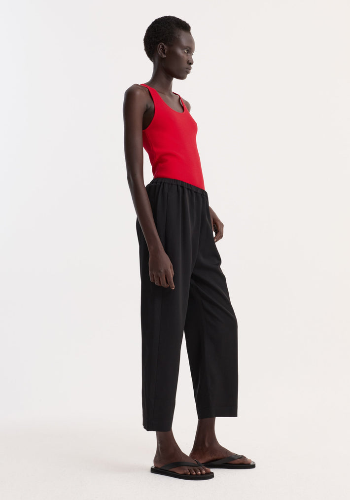 Model in profile wearing Cropped wide leg trousers | black, showcasing their loose fit, ankle length, elastic waistband, and side pockets. Paired with a red tank top.
