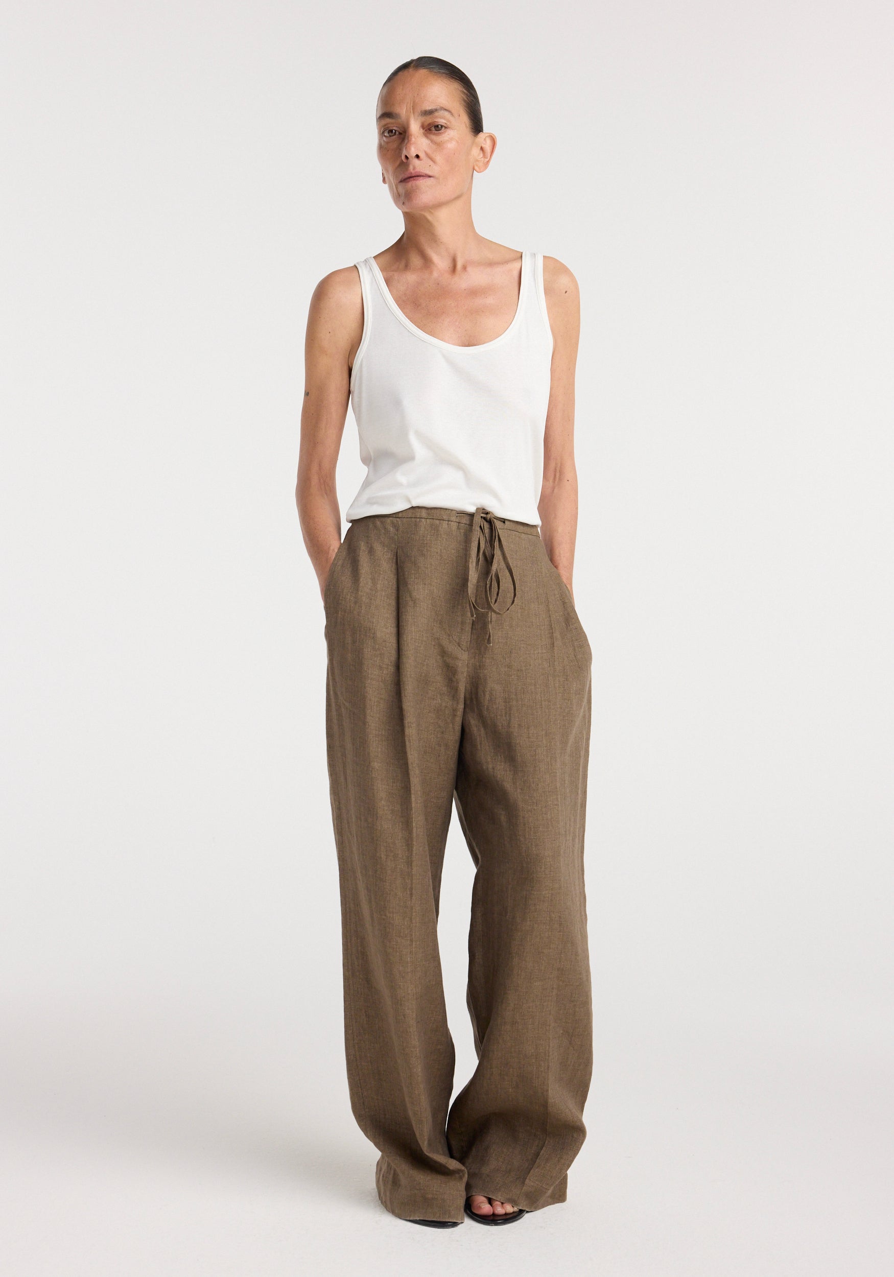 Woman wearing relaxed drawstring trousers | brown melange paired with a white sleeveless top, standing against a plain light background with hands in pockets.