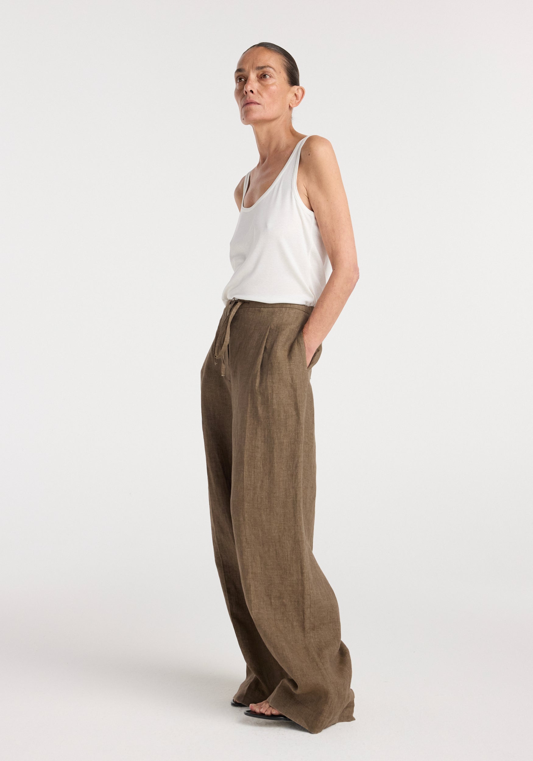 Woman wearing relaxed drawstring trousers | brown melange with wide legs and a high waist, paired with a white sleeveless top against a plain background.
