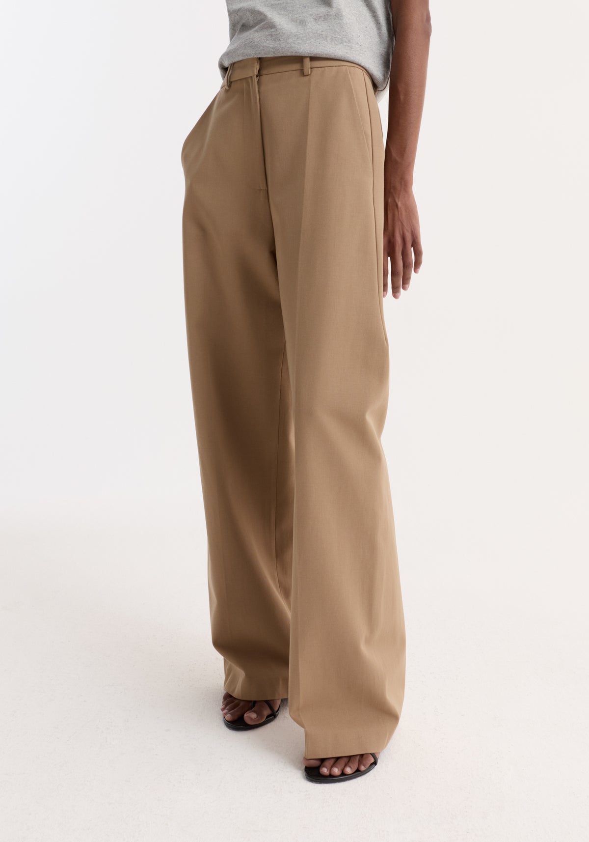 A person wearing Classic loose fit chino | beige pants paired with a tucked-in grey top and black open-toe shoes, standing against a plain white background.