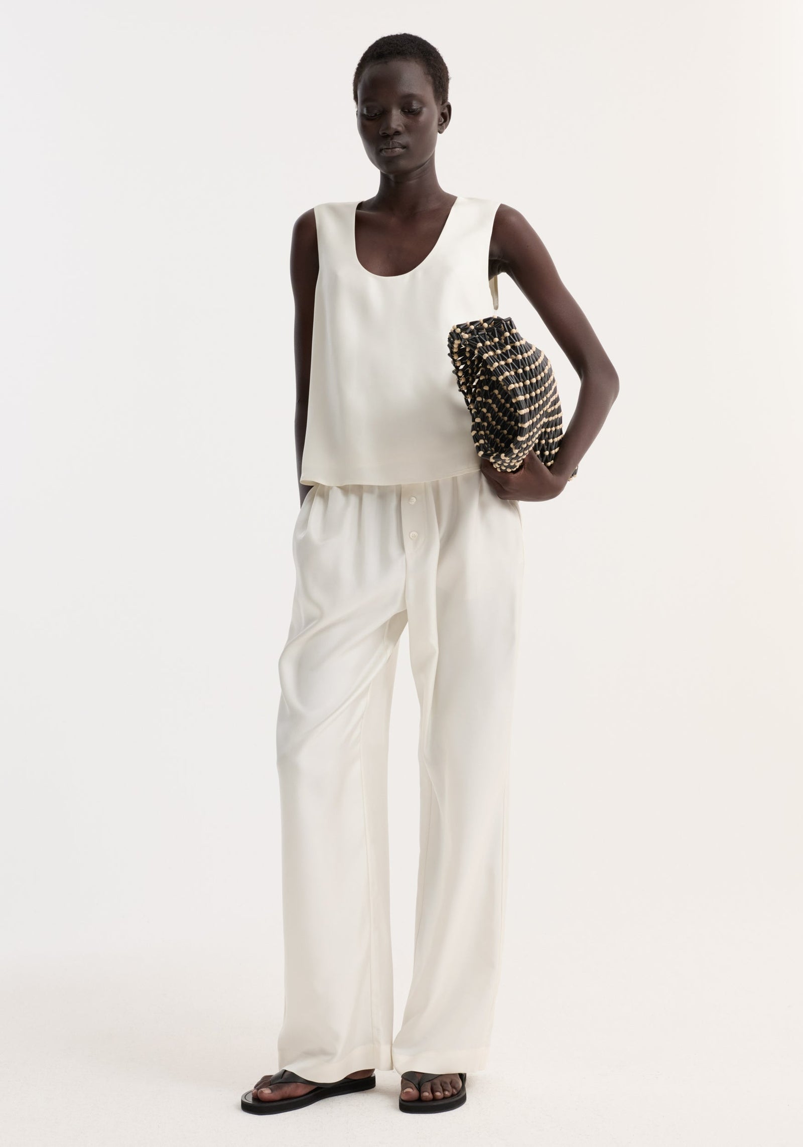 Model wearing Silk pyjama trousers | cream, with a sleeveless top and holding a textured clutch. The outfit is minimalist, with contrast from the woven bag.