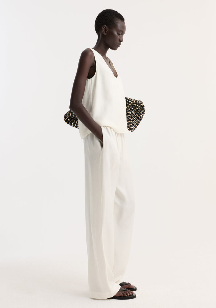 Model in profile wearing Silk pyjama trousers, paired with a sleeveless top, holding a woven clutch, and wearing black thong sandals.