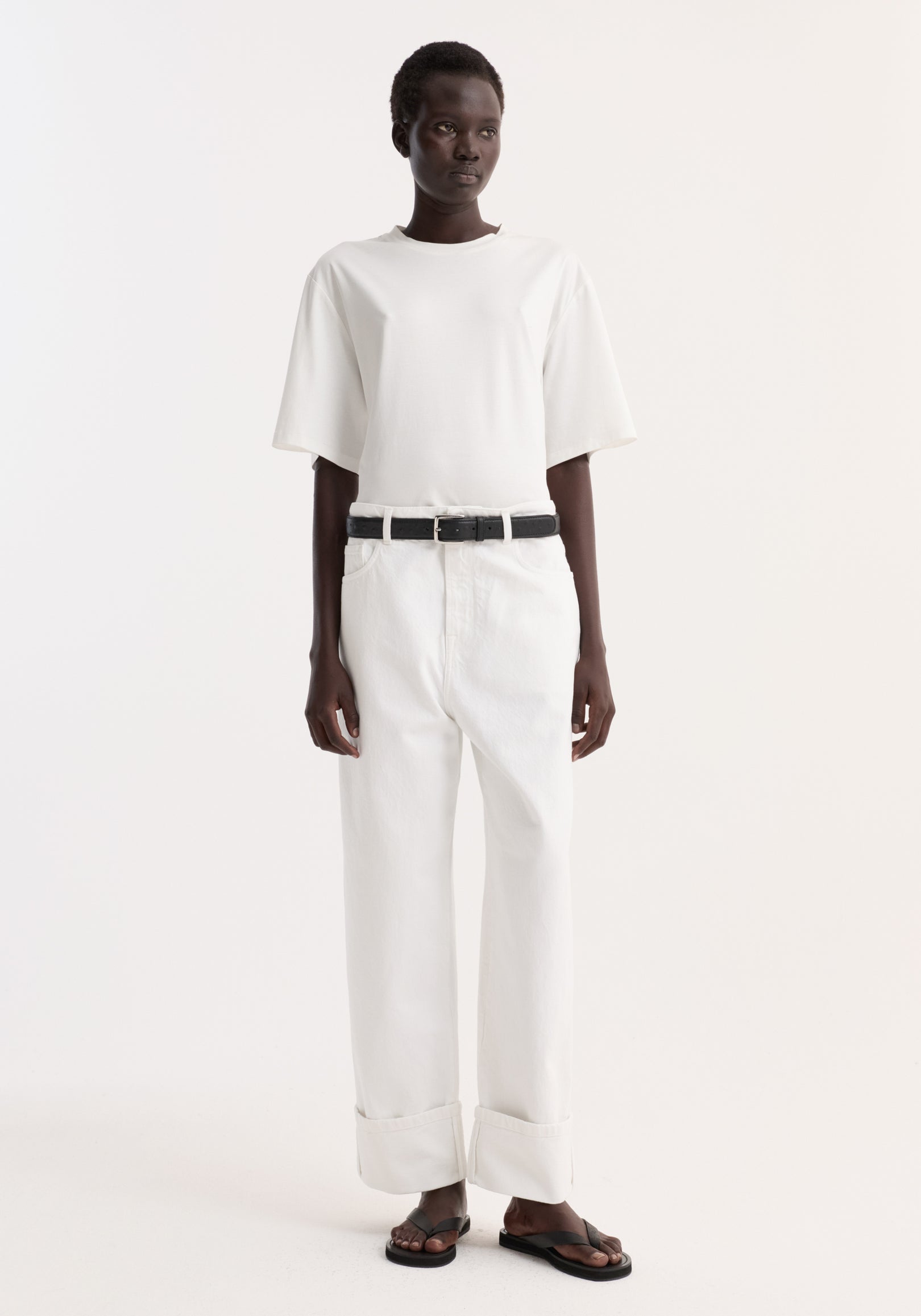 Model wearing Baggy fit turn-up denim in optic white, showcasing low-waist, baggy, wide-leg style with turned-up hems, paired with a white t-shirt and black sandals.