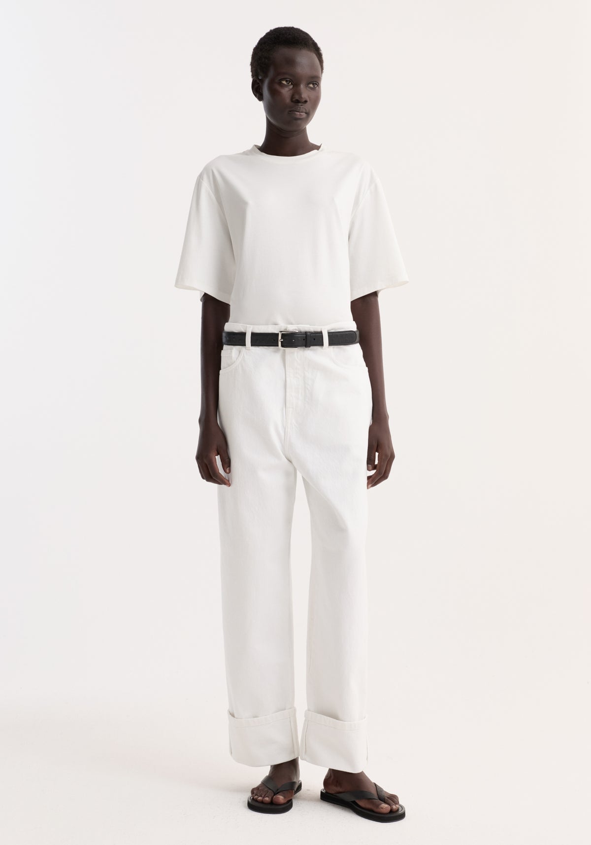 Model wearing Baggy fit turn-up denim in optic white, showcasing low-waist, baggy, wide-leg style with turned-up hems, paired with a white t-shirt and black sandals.