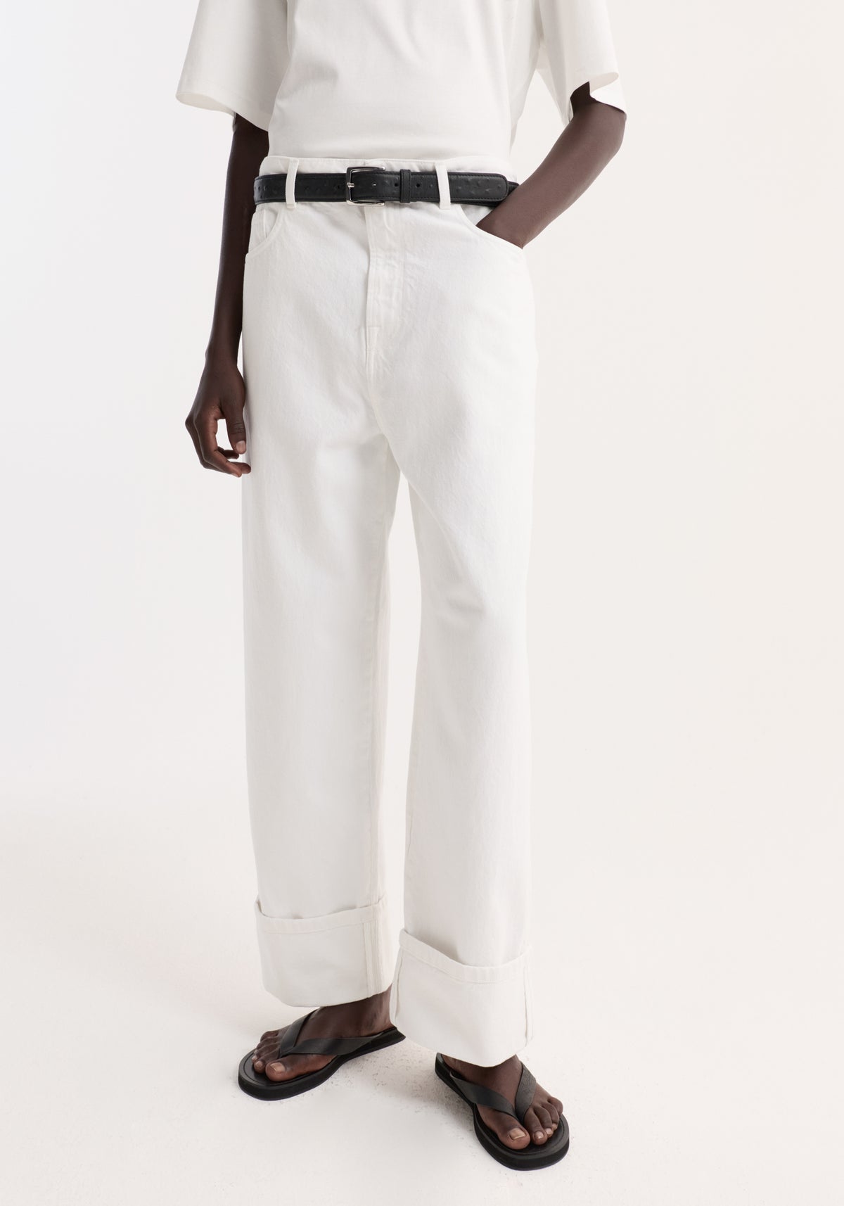 Baggy fit turn-up denim in optic white, featuring a wide-leg, low-rise style. Modeled with a black belt and flip-flop sandals, emphasizing casual elegance.