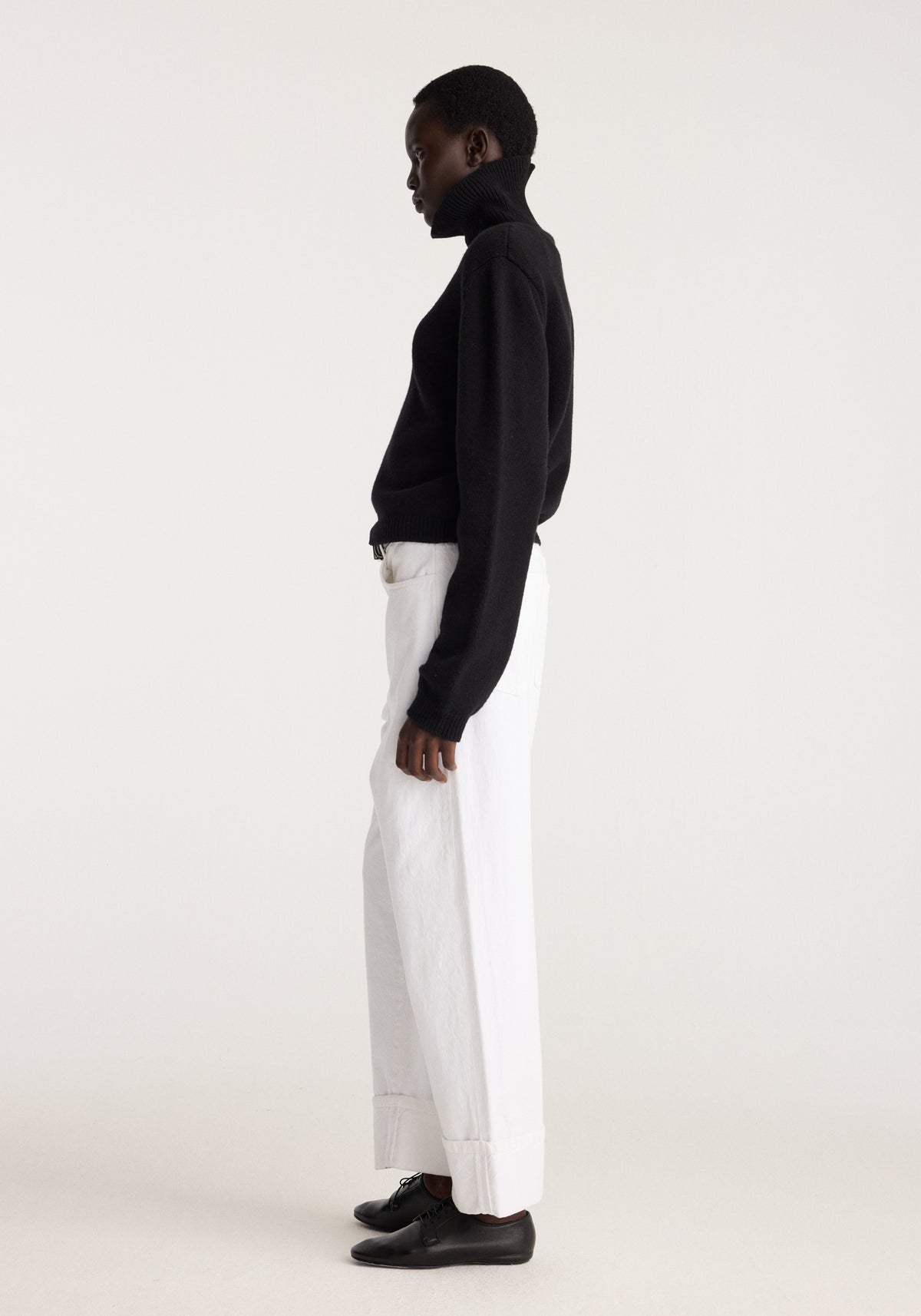 Side view of a model wearing Baggy fit turn-up denim | optic white with a black sweater and black shoes against a light studio background