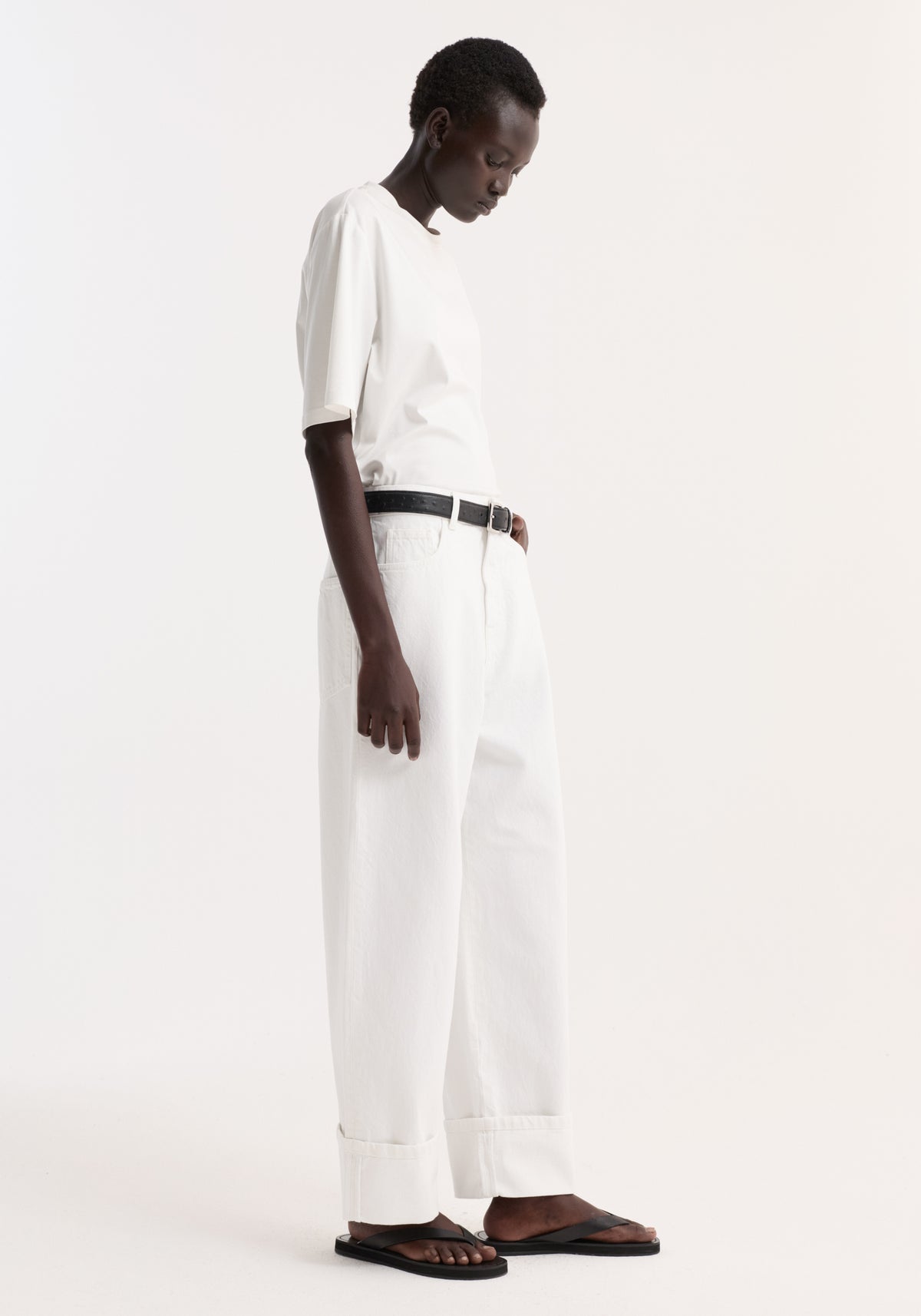 Woman in profile wearing Baggy fit turn-up denim in optic white, paired with a white T-shirt, black belt, and black thong sandals.