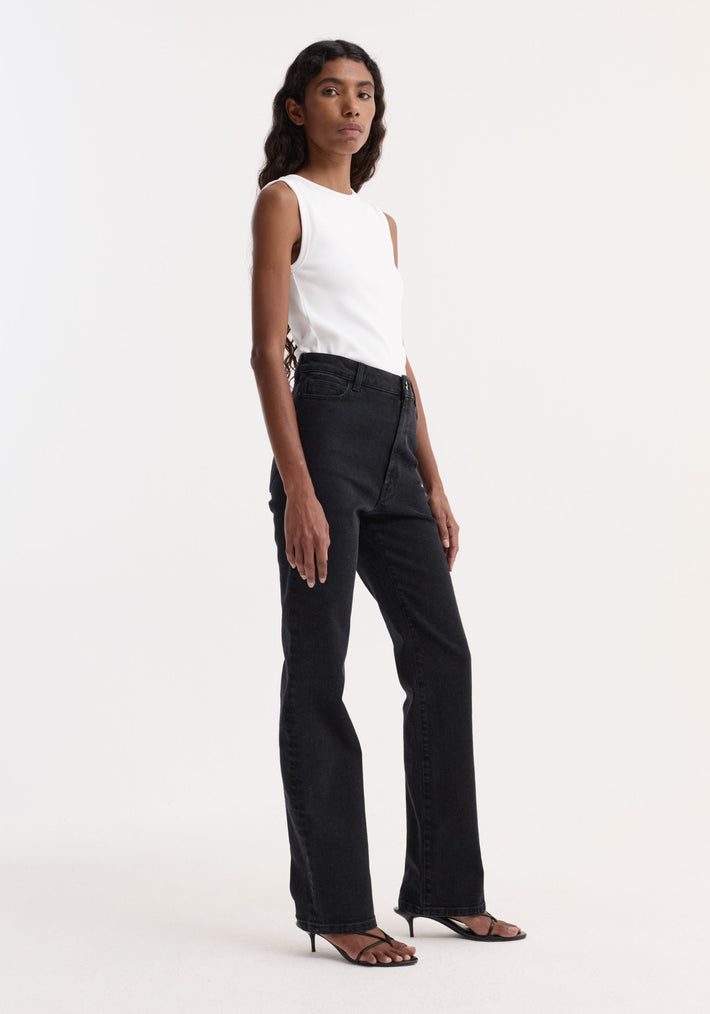 Woman wearing high-rise straight stretch denim in black, featuring a slim leg and higher waist, paired with a sleeveless top and heeled sandals.