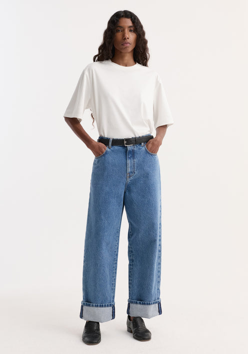 Person wearing Turn-up baggy denim | heritage blue with rolled cuffs, a white T-shirt, black belt, and black slip-on shoes, hands in pockets.