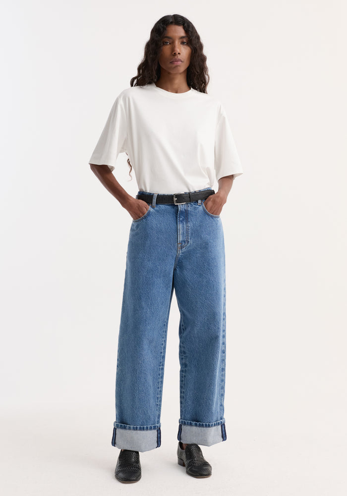Person wearing Turn-up baggy denim | heritage blue with rolled cuffs, a white T-shirt, black belt, and black slip-on shoes, hands in pockets.