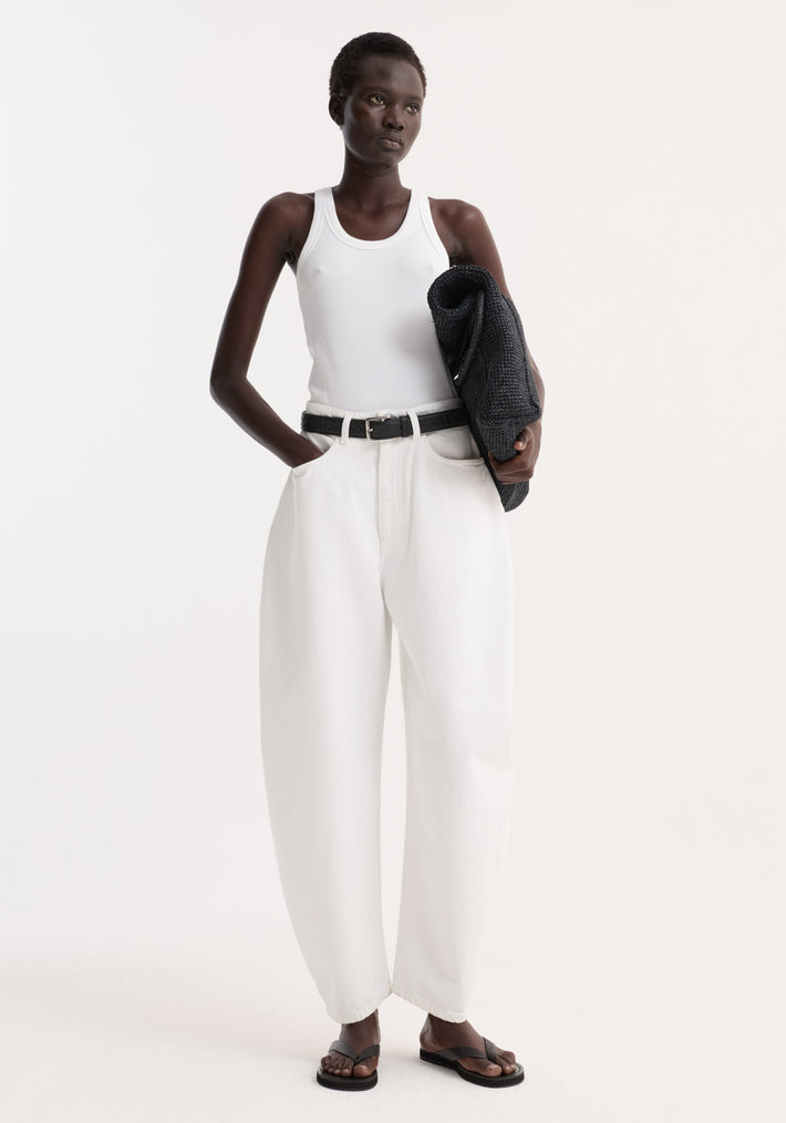 Model in optic white high-waist sculptural denim with balloon leg, paired with a white tank top and black sandals, holding a dark knit sweater.