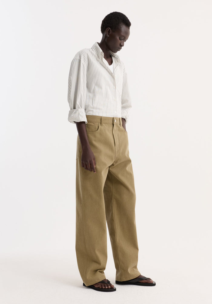Model in Baggy fit denim | sage, wearing oversized striped shirt, white tank, camel-wide-leg trousers, black sandals, posed with hand in pocket against white background.