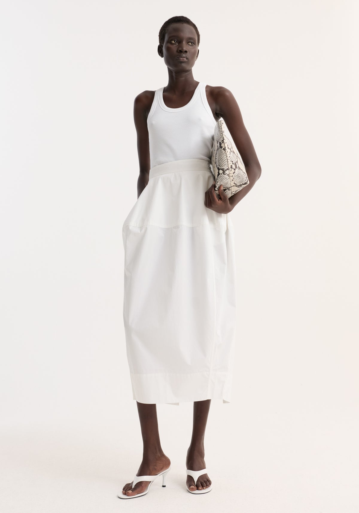 Woman wearing Sculptural poplin skirt in optic white with high waist, seam detailing, and back slit, paired with a white tank top and heeled sandals.