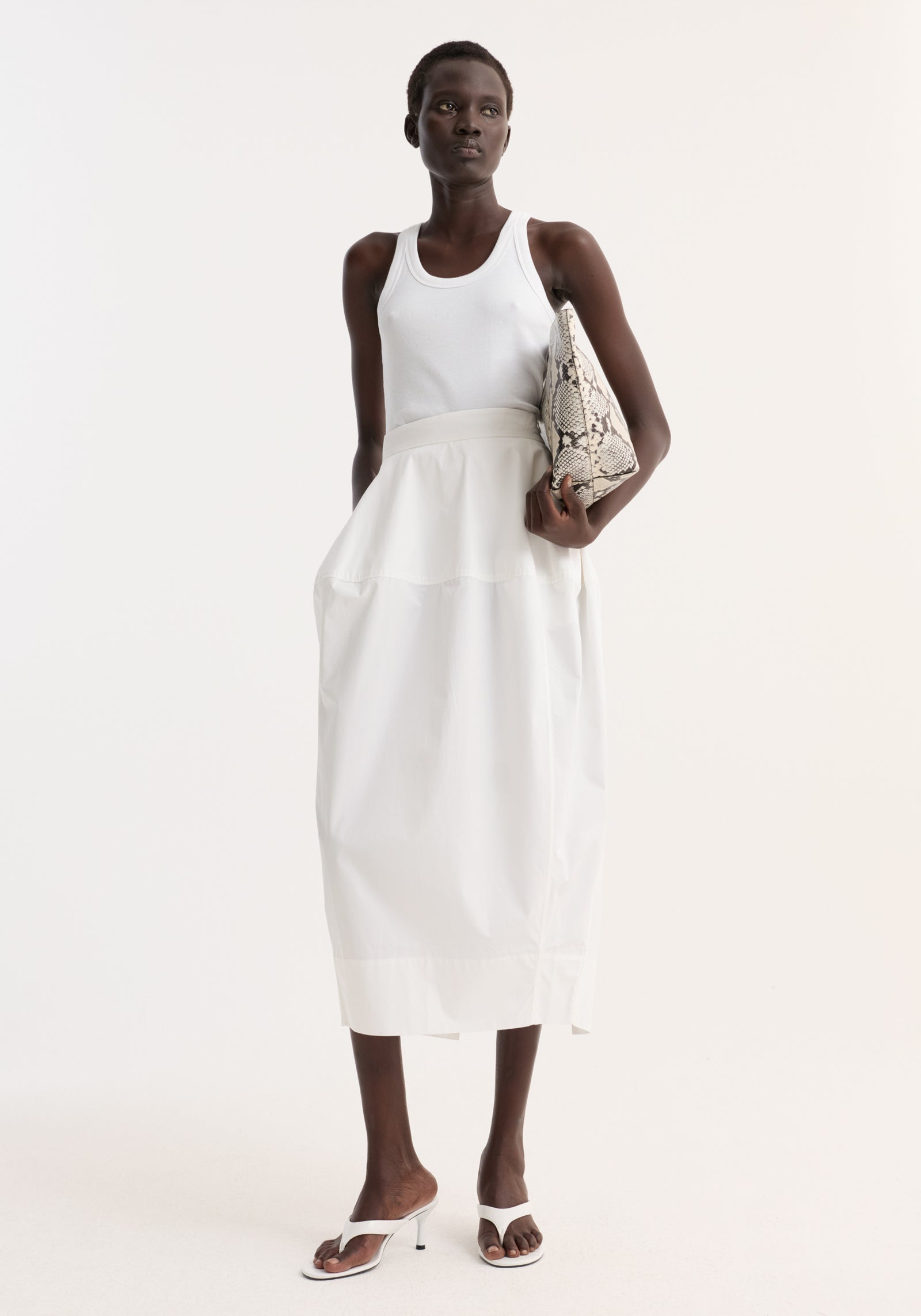 Woman wearing Sculptural poplin skirt in optic white with high waist, seam detailing, and back slit, paired with a white tank top and heeled sandals.