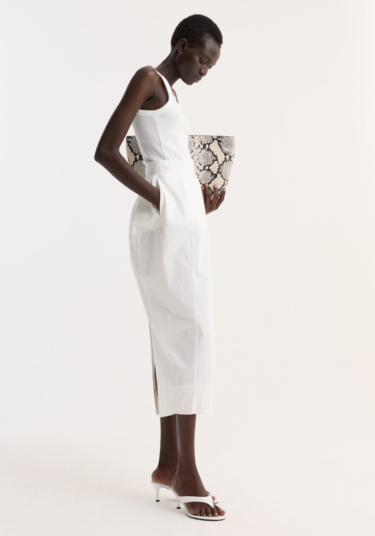 Model in profile wearing Sculptural poplin skirt in optic white, featuring a high waist and seam detailing, styled with a white tank top and python-print tote.