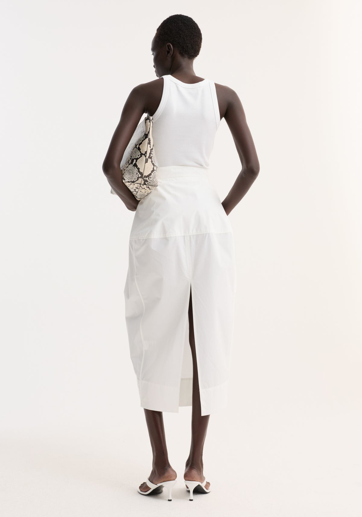Woman in a white sculptural poplin skirt with high waist and back slit, paired with a white rib-tank top and snakeskin print clutch.