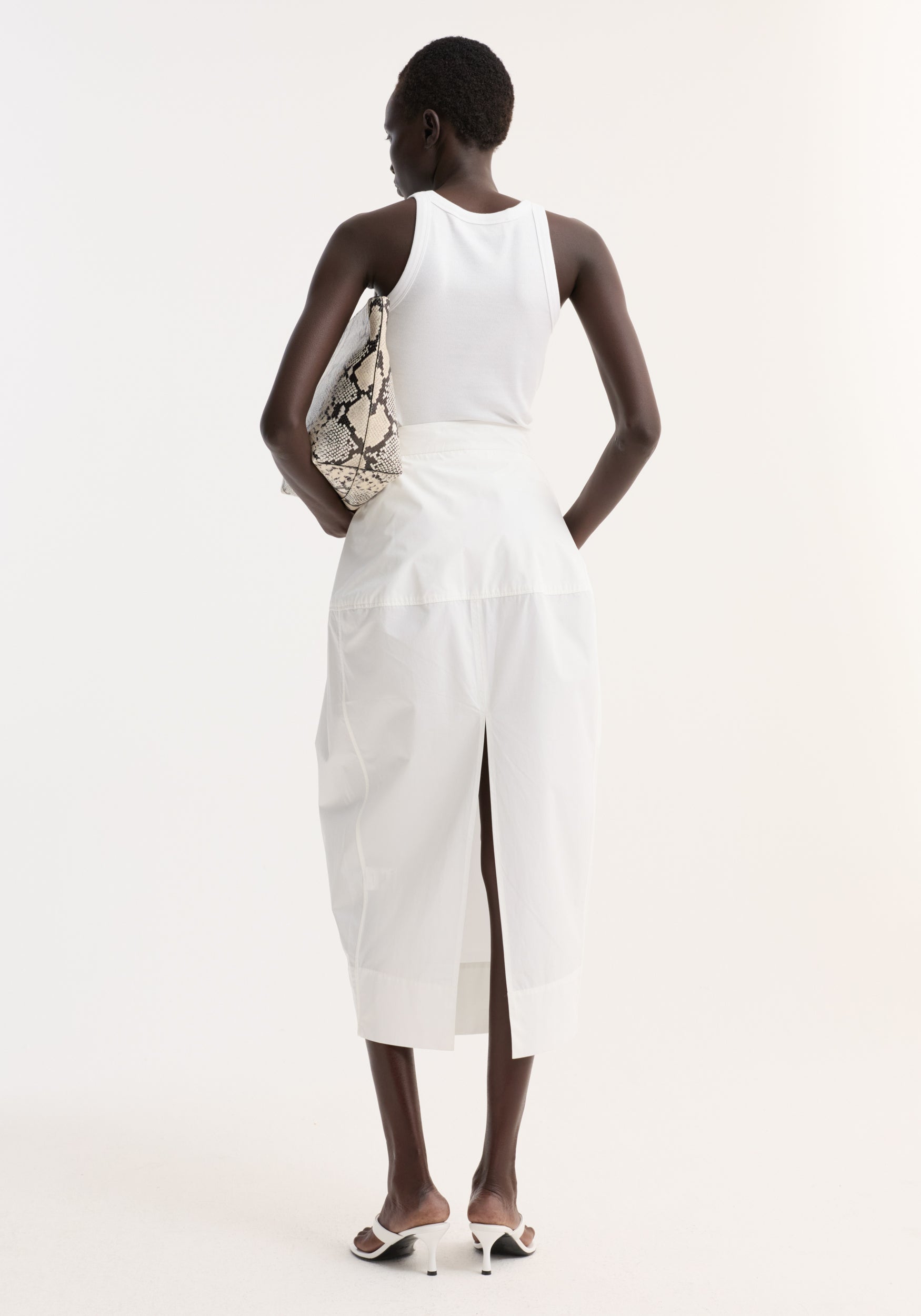 Woman in a white sculptural poplin skirt with high waist and back slit, paired with a white rib-tank top and snakeskin print clutch.