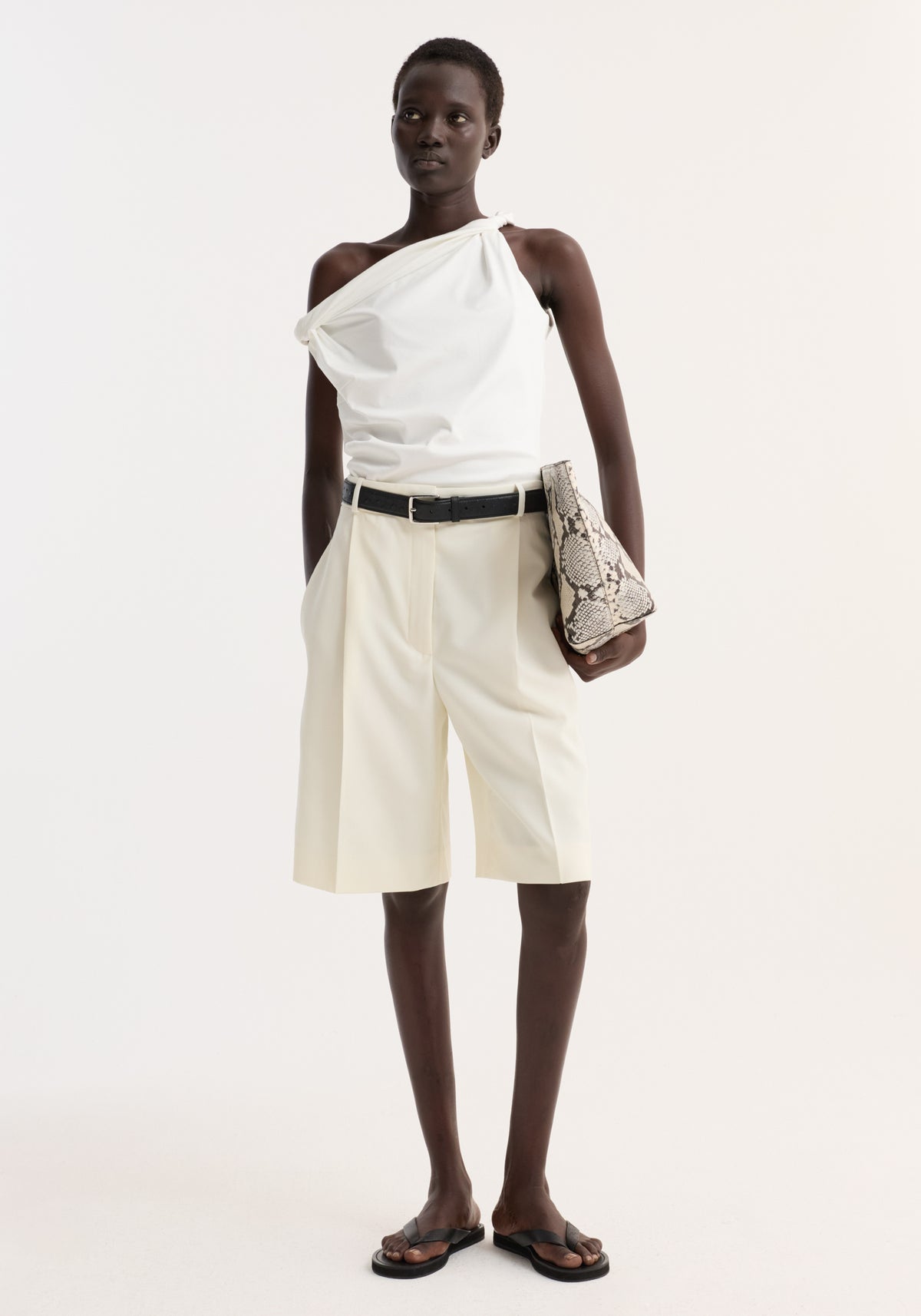Model wearing tailored single pleat shorts | off white paired with a white asymmetrical sleeveless top, black belt, black sandals, and holding a snakeskin-patterned clutch bag against a plain background.