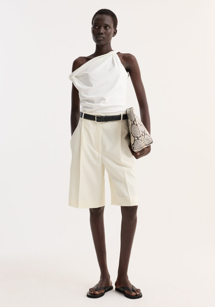 Model in Tailored single pleat shorts | off white, high-waisted with side pockets, paired with a one-shoulder top and black accessories, holding a snakeskin clutch.