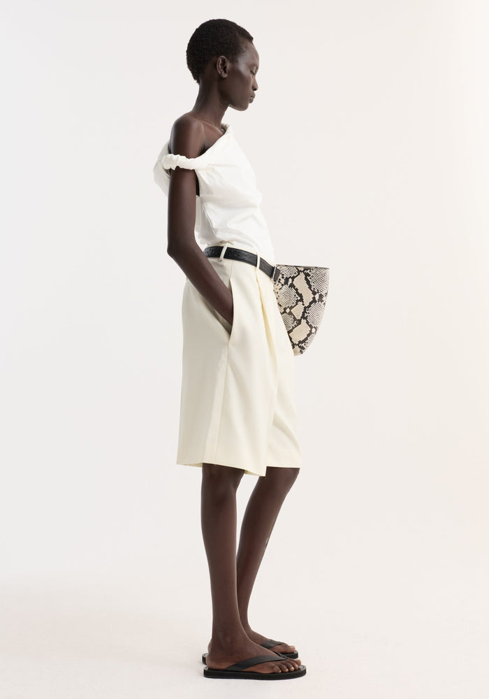 Model wearing tailored single pleat shorts in off white with side pockets and a front pleat, styled with a black belt and flat sandals.