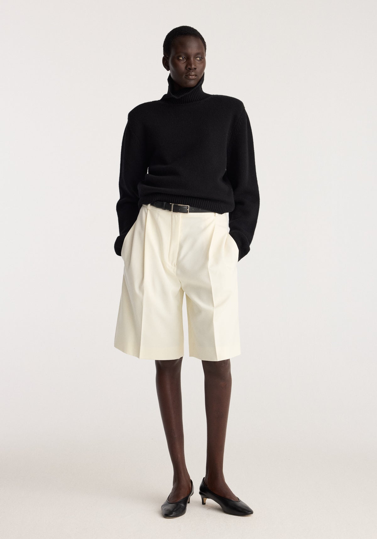 Tailored single pleat shorts | off white styled with a black turtleneck sweater and black shoes on a model in a neutral studio setting
