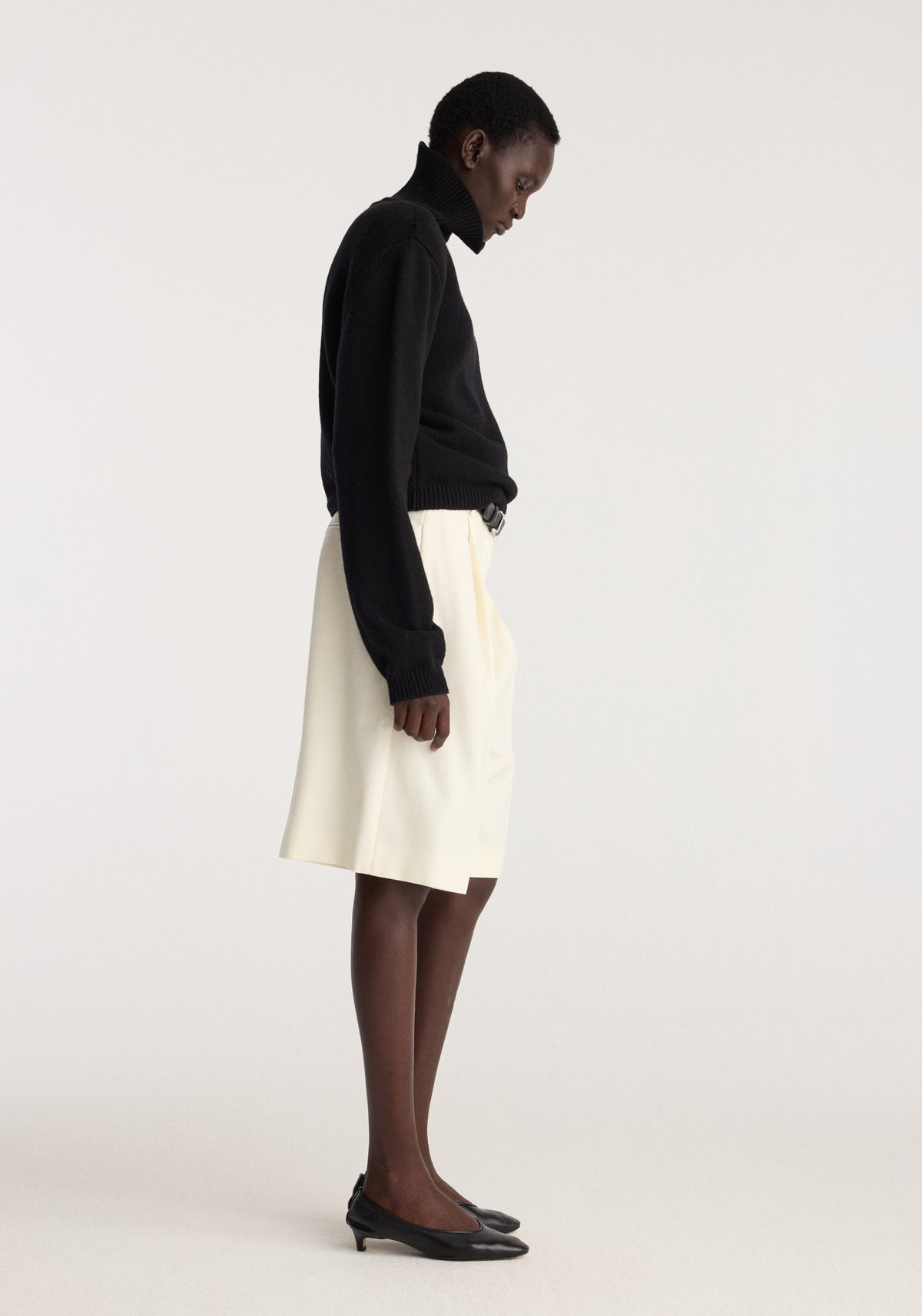 Tailored single pleat shorts | off white shown in a side profile outfit with a black sweater and black pointed flats in a studio setting