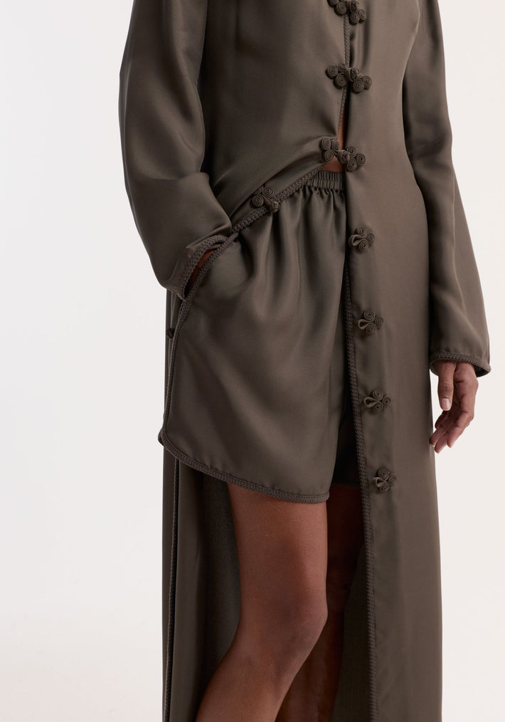 Person wearing dark olive-brown coat with Taped silk shorts | earth, featuring an elastic waistband, pleats, and embroidered detailing. Hand rests in pocket.