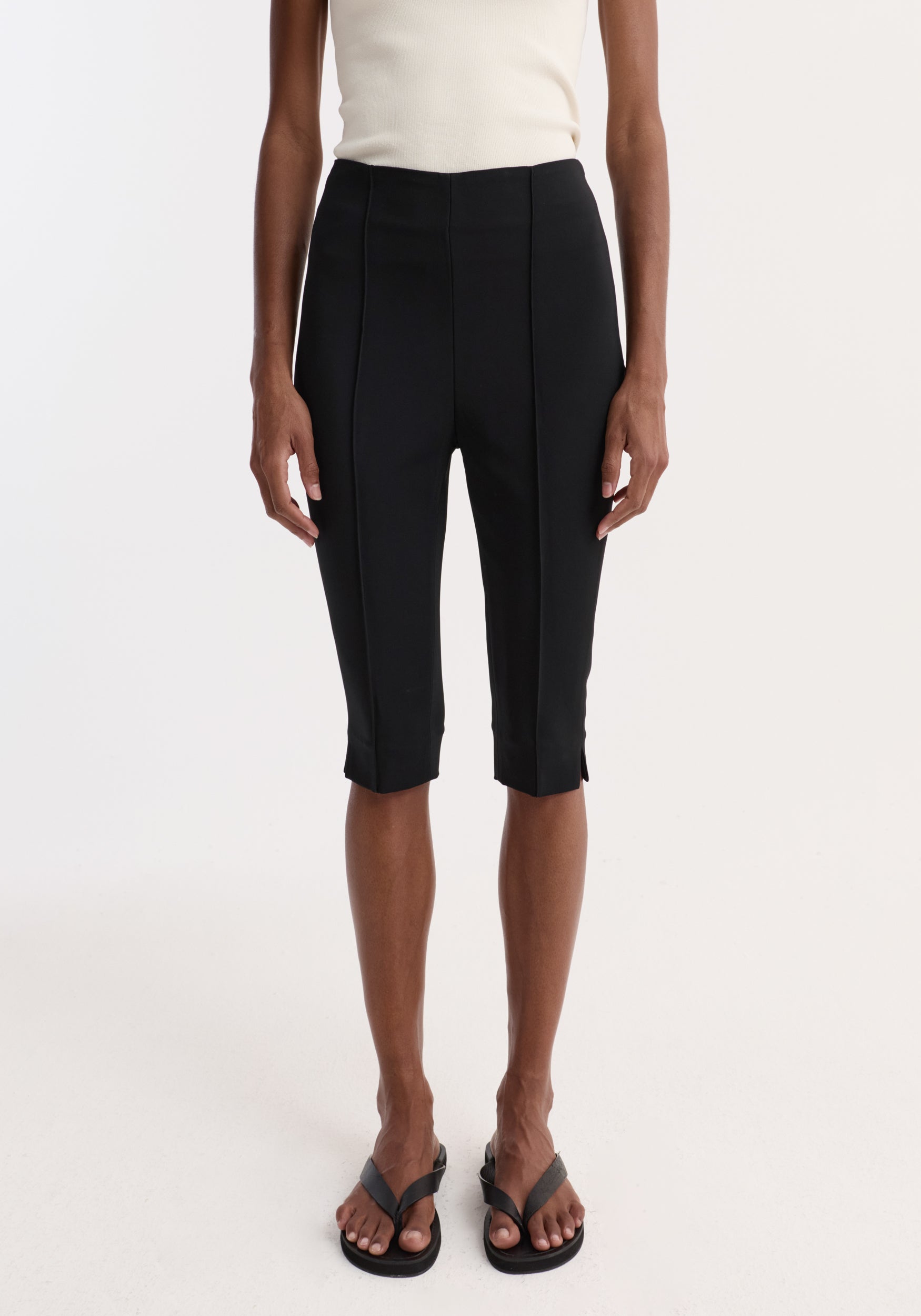 Front view of a person wearing Capri | black fitted knee-length pants paired with black sandals and a light-colored sleeveless top against a plain white background.