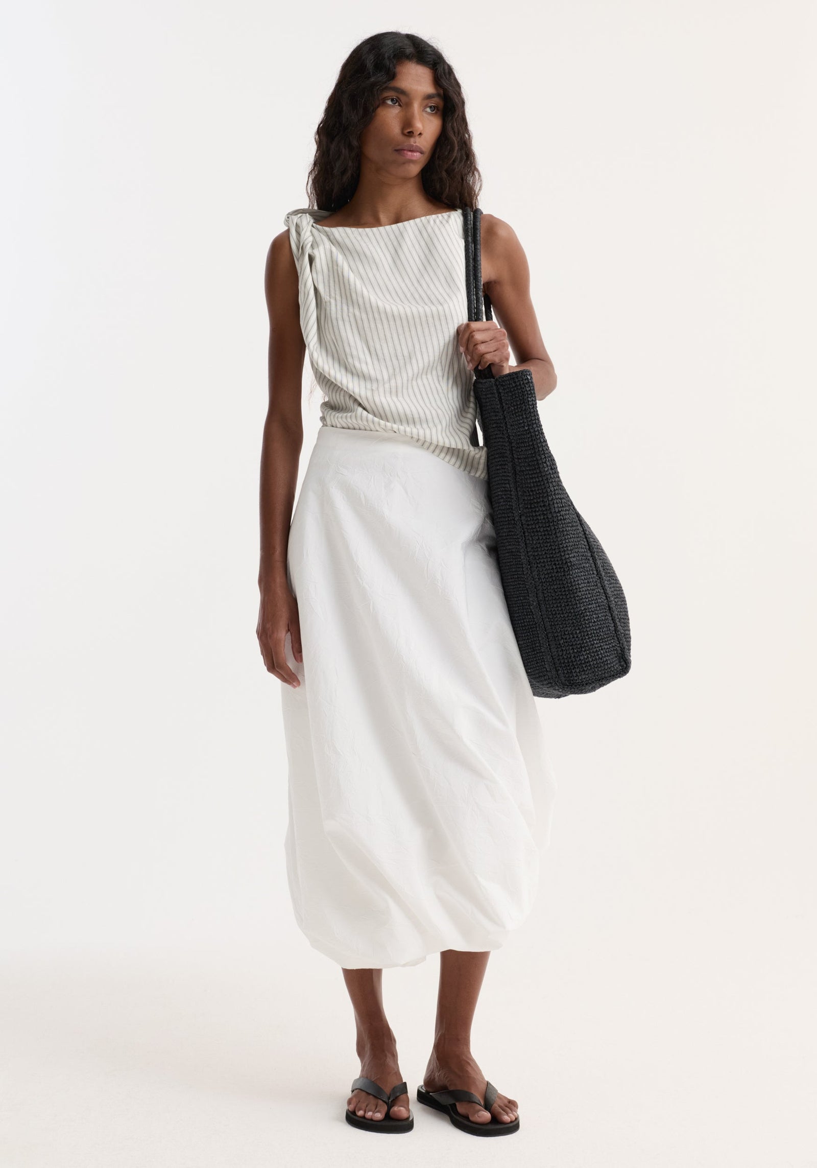 Model wearing Twisted crushed cotton skirt | optic white, featuring high-waisted, voluminous balloon hem, paired with a pinstriped top, black tote, and sandals.