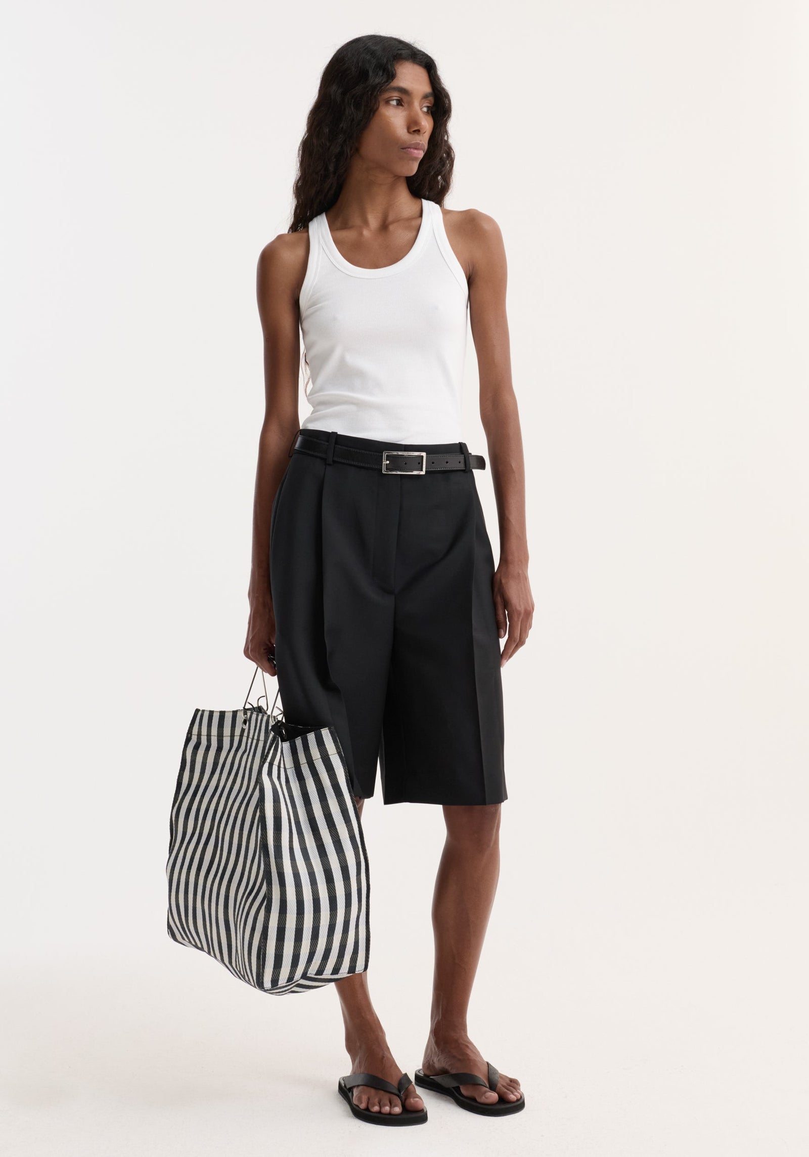 Model wearing tailored single pleat shorts, black, with side pockets and knee-length cut, styled with a white tank top, black belt, sandals, and striped tote.
