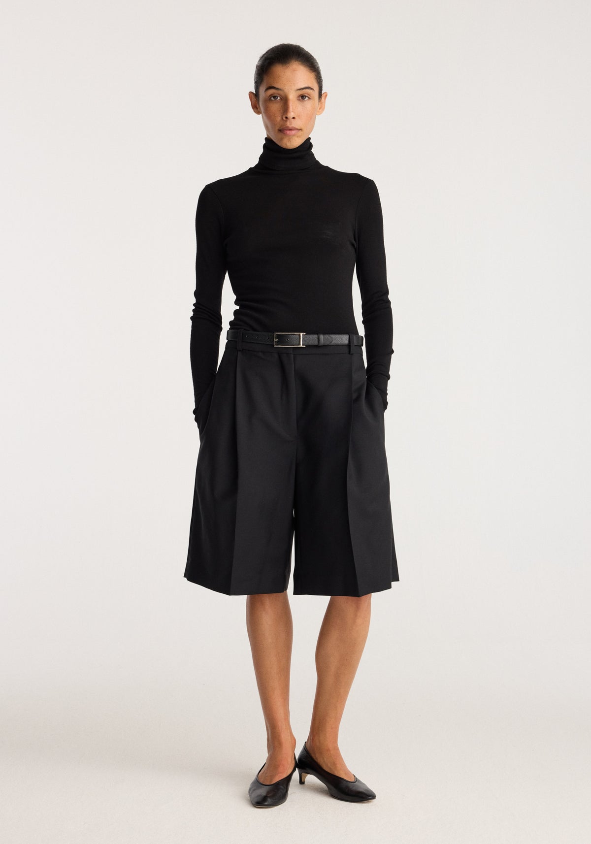 Tailored single pleat shorts | black worn by a model with a black turtleneck and belt in a studio setting